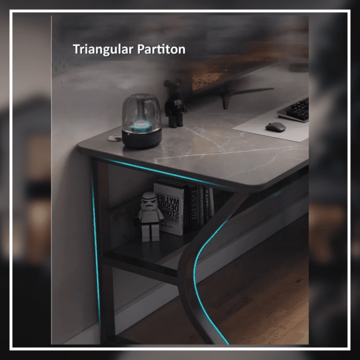 A modern gaming desk setup featuring a sleek, gray desk with a curved black frame. The desk holds a monitor displaying a mountain landscape, a keyboard, a mouse, and various accessories. A gaming PC with blue LED lights is positioned underneath. The wall has a pegboard with a digital clock and other items.