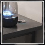 A modern gaming desk setup featuring a sleek, gray desk with a curved black frame. The desk holds a monitor displaying a mountain landscape, a keyboard, a mouse, and various accessories. A gaming PC with blue LED lights is positioned underneath. The wall has a pegboard with a digital clock and other items.