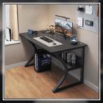 A modern gaming desk setup featuring a sleek, gray desk with a curved black frame. The desk holds a monitor displaying a mountain landscape, a keyboard, a mouse, and various accessories. A gaming PC with blue LED lights is positioned underneath. The wall has a pegboard with a digital clock and other items.