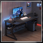 A modern gaming desk setup featuring a sleek, gray desk with a curved black frame. The desk holds a monitor displaying a mountain landscape, a keyboard, a mouse, and various accessories. A gaming PC with blue LED lights is positioned underneath. The wall has a pegboard with a digital clock and other items.